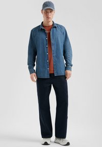 Jeans overhemd over een roestkleurig T-shirt, gecombineerd met een marineblauwe broek en grijze sneakers. Een blauwe pet maakt de outfit compleet.