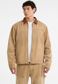 Giacca di cotone beige con collo in corduroy marrone, zip frontale e tasche laterali. Presenta dettagli di cucitura discreti e una vestibilità comoda.