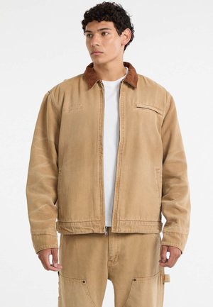 Tan cotton jacket with a brown corduroy collar, front zipper, and side pockets. Features subtle stitching details and a relaxed fit.