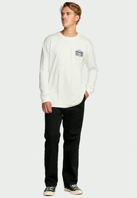 Jonge man staat, draagt een wit shirt met lange mouwen en een kleine zwarte grafische afbeelding op de borst, zwarte broek en zwarte sneakers met witte zolen.