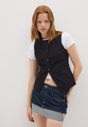 Gilet noir ajusté à col haut, avec des boutons à l'avant et un ourlet pointu, porté sur un t-shirt blanc et associé à une mini-jupe en denim.