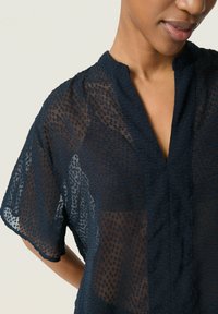 Blusa blu navy realizzata in un tessuto leggero e trasparente con un motivo testurizzato, maniche corte e scollo a V con bordo arricciato.