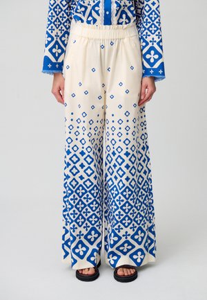 Pantalones de pierna ancha en tejido crema con patrones geométricos azules. Presentan una cinturilla elástica y una textura sutil, combinados con sandalias negras.