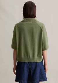 Zielony sweter wykonany z wełny z domieszką, z kołnierzykiem, krótkimi rękawami i ściągaczem, w połączeniu z ciemną dżinsową spódnicą z plisowanym wzorem.