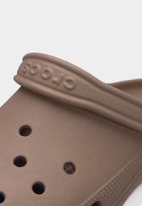 Καφέ clog Crocs με οπές αερισμού και ρυθμιζόμενο λουράκι φτέρνας με ανάγλυφο το όνομα της μάρκας.