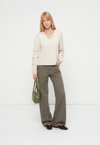 Beige v-kaula-aukollinen paita, jossa on liehuvat hihat, yhdistetty oliivinvihreisiin leveälahkeisiin housuihin. Mallilla on pieni vihreä käsilaukku ja päällä viininpunaiset kengät.