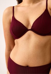 Donna che indossa un reggiseno e slip coordinati in maglia costina color amaranto, mostrando la parte superiore del torso e la zona centrale contro uno sfondo neutro.