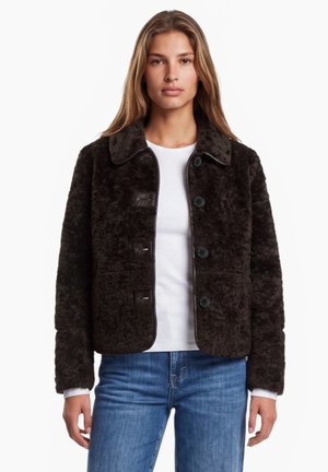 Femme portant une veste boutonnée marron foncé duveteuse par-dessus une chemise blanche et un jean bleu, debout devant un fond blanc uni.