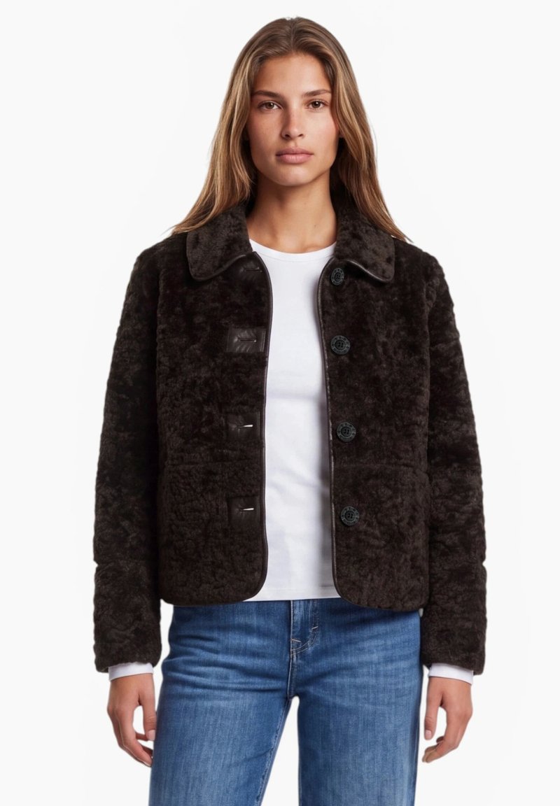 Femme portant une veste boutonnée marron foncé duveteuse par-dessus une chemise blanche et un jean bleu, debout devant un fond blanc uni.