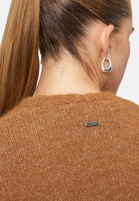 Pull marron en tricot avec une texture douce, doté d'une petite étiquette en métal à l'arrière. Le modèle a les cheveux longs attachés en arrière et porte des boucles d'oreilles argentées.