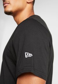 Hombre vistiendo una camisa de manga corta negra con el logo blanco de New Era bordado en la manga izquierda sobre un fondo liso.