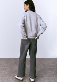 Sudadera gris con puños acanalados, combinada con pantalones vaqueros anchos de color gris oscuro con un dobladillo deshilachado. Zapatillas de color claro completan el atuendo.