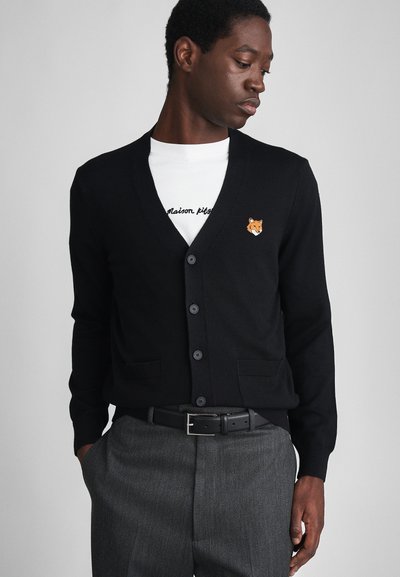 Cárdigan negro con diseño de pico, que presenta detalles bordados de un zorro en el pecho, llevado sobre una camisa blanca con texto, combinado con pantalones grises.