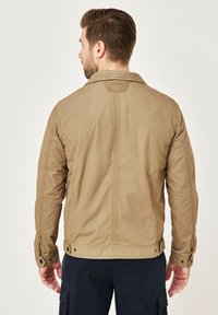 Brown cotton jacket with a plain design, buttoned cuffs, and a straight cut. Features a back yoke and visible stitching details.