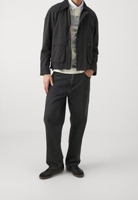 Mand iført en mørk jakke over en stribet grå og hvid polo, sorte løse jeans og sorte sneakers foran en ensfarvet baggrund.