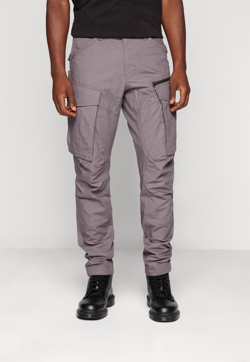 Homme portant un pantalon cargo gris avec plusieurs poches et des bottes noires lacées, debout devant un fond clair uni.