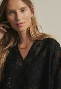 Sort gennemsigtig bluse med blomsterbroderi, struktureret stof og en bølget halsudskæring. Modellen bærer et guld snoet kædeneklæde.