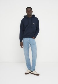 Φούτερ σε σκούρο μπλε χρώμα με ρυθμιζόμενα κορδόνια και μπροστινή τσέπη, συνδυασμένο με ανοιχτό μπλε τζιν και αθλητικά παπούτσια σε εκρού χρώμα. Απλός σχεδιασμός.