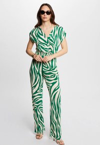 Grøn og beige zebra-stribet jumpsuit med korte ærmer, elastisk talje og flagrende ben, parret med sorte solbriller og guldfarvede accessories.
