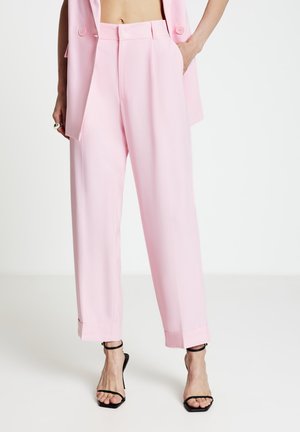 Pantalon rose sur mesure à coupe ample, avec une taille haute, des plis devant, des poches latérales et des revers roulés. Associé à des escarpins noirs à fines lanières.