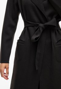 Cappotto nero avvolgente realizzato in tessuto liscio, con cintura annodata, due tasche frontali e maniche lunghe. Design minimalista senza motivi visibili.