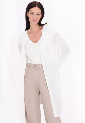Cardigan bianco leggero in maglia con apertura frontale e maniche ampie, indossato sopra un top bianco e pantaloni beige. Texture morbida, design casual.