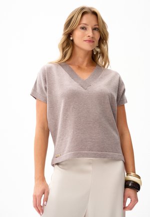 Kobieta ubrana w taupe krótkawy rękaw, sweter z dekoltem w serek, kremowe spodnie oraz nałożone na prawe nadgarstek bransoletki na białym tle.