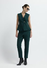 Gilet peplum e pantaloni aderenti in tessuto verde scuro. Il gilet presenta un profondo scollo a V e un singolo bottone dorato. La modella indossa degli stivaletti neri alla caviglia.