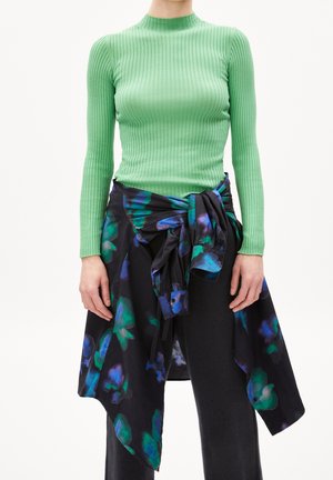 Groene geribbelde top met lange mouwen gecombineerd met een zwarte wikkel met bloemenpatroon, met groene en blauwe accenten en een geknoopte detail bij de taille.