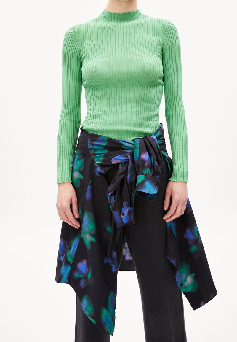 Grön ribbad långärmad topp tillsammans med en svart blommönstrad omlott, med gröna och blå accenter samt en knuten detalj vid midjan.