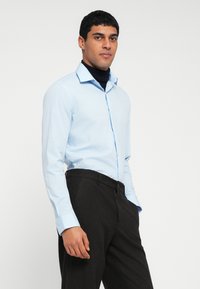 Hele sinine pikkade varrukatega särk terava kraega, ees nööbitav, kantud tumedate pidulike pükstega, luues kontrasti värvi ja tekstuuri vahel.