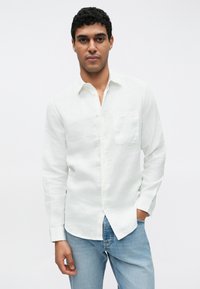 Camisa de linho branca com mangas compridas, fecho de botões na frente e um bolso no peito, combinada com jeans de ganga azul claro.