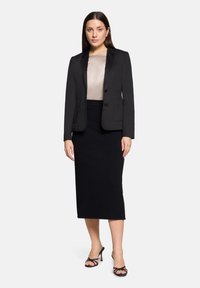 Femme portant un blazer noir, un chemisier beige, une jupe midi noire et des sandales à talons hauts noires, debout devant un fond clair uni.