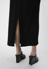 Robe maxi noire avec une fente au dos, associée à des mules à talons épais noires. Tissu lisse avec une texture élégante et un design minimaliste.