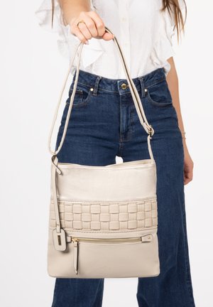 Frau hält beige geflochtene Lederschultertasche mit Reißverschlusstasche vorne, trägt weiße Bluse und blaue Jeans.