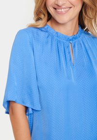 Blusa azzurra a maniche corte realizzata in tessuto leggero con un sottile motivo a pois, caratterizzata da un collo arricciato e un dettaglio a buco della serratura.