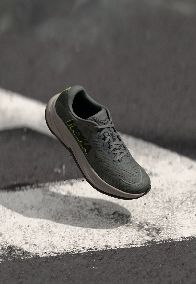 Sapatilha de corrida cinza com um revestimento texturizado, detalhes em verde e sola leve, mostrada no ar acima de uma superfície escura com marcas brancas.