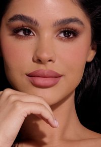 Zbliżenie twarzy o gładkiej, jasnobrązowej skórze, pełnych matowych w kolorze mauve ustach, wyrazistych rzęsach i wyraźnych brwiach, w naturalnym makijażu.