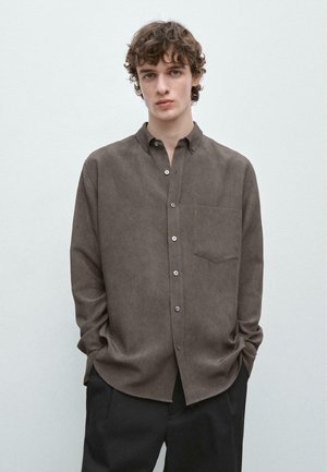 Junger Mann mit kurzen lockigen Haaren, trägt ein lockeres braunes Hemd mit Knöpfen und Brusttasche sowie schwarze Hose, Hände in den Taschen, neutraler Hintergrund.