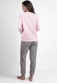Mujer de pie descalza, vistiendo una blusa de manga larga rosa y pantalones de estampado de leopardo, contra un fondo blanco liso, de espaldas.