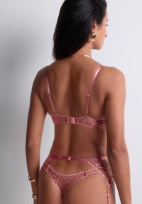 Femme aux longs cheveux noirs portant de la lingerie en dentelle rose, des boucles d'oreilles en or et des bijoux en or, vue de dos sur un fond blanc uni.