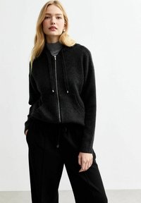 Zwarte hoodie met ritssluiting, gemaakt van zachte stof met een gestructureerde afwerking. Voorzien van trekkoorden, verlaagde schouders en een losse pasvorm. Gecombineerd met zwarte broek.
