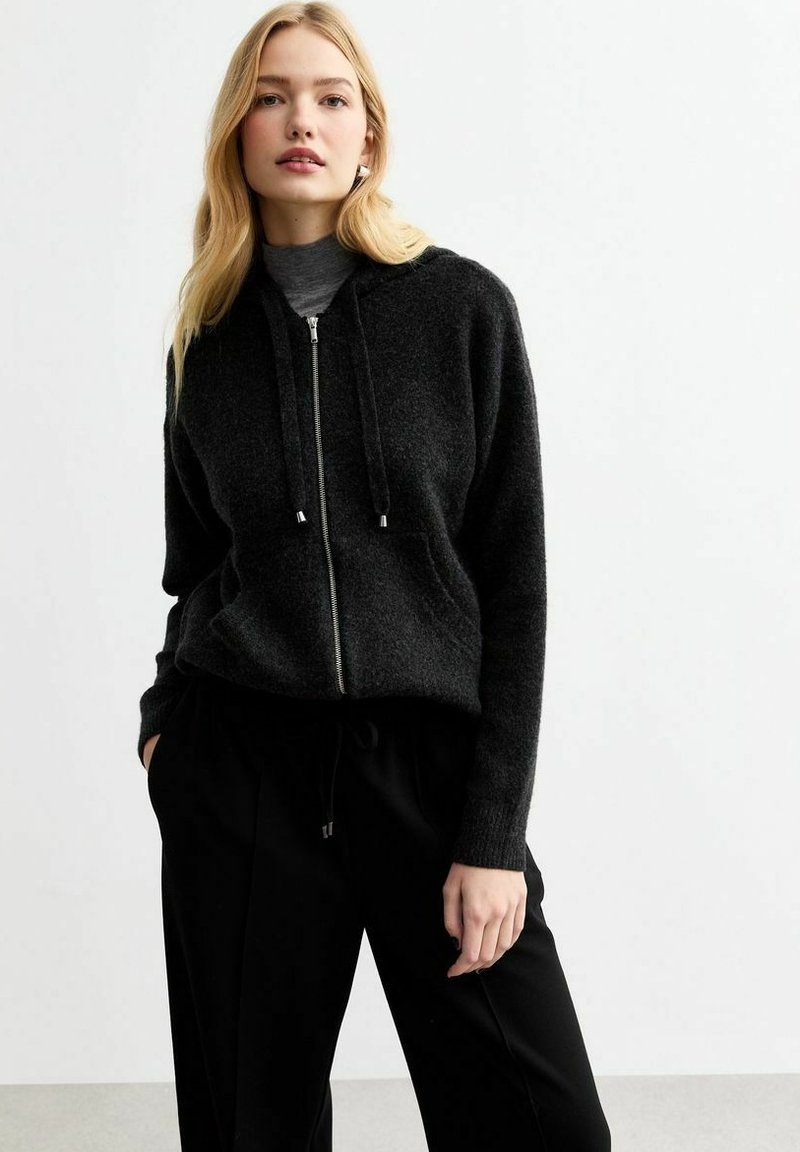 Zwarte hoodie met ritssluiting, gemaakt van zachte stof met een gestructureerde afwerking. Voorzien van trekkoorden, verlaagde schouders en een losse pasvorm. Gecombineerd met zwarte broek.