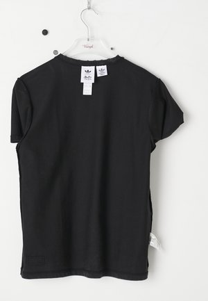 Czarna koszulka z krótkim rękawem wisząca na przezroczystym wieszaku Triumph, pokazująca tylną stronę oraz trzy białe metki Adidas i Buffy.