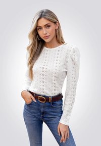 Witte lange mouwen top met een floral kantpatroon, ronde halslijn en opgestroopte schouders, gecombineerd met blauwe jeans en een bruine riem.