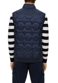 Navy-blaues gepolstertes West mit abgerundetem gestepptem Design, hohem Kragen und ohne sichtbare Hardware, darüber getragen ein gestreiftes Langarmshirt.