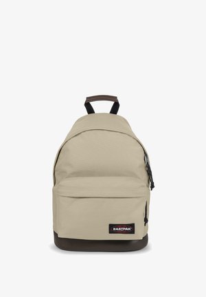 Beige Rucksack mit runder Form, strukturiertem Stoff, braunem Boden, Fronttasche und schwarzen Trägern. Verfügt über ein Logopatch an der Vorderseite.