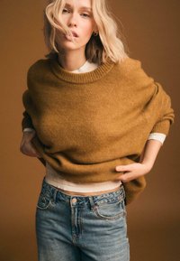 Hořčicově zbarvený oversized svetr s žebrovaným výstřihem, nošený přes bílý top, doplněný o světlé modré džíny, na hnědém pozadí.