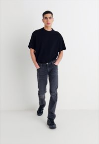 Jeune homme marchant en avant, les mains dans les poches, portant un t-shirt noir surdimensionné, un jean sombre et des sneakers noires sur un fond blanc.