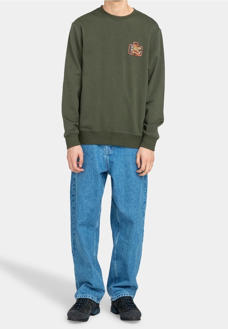 Groen sweatshirt met ronde hals en geribbelde boorden, voorzien van een klein logoplakje. Gecombineerd met lichtblauwe, losvallende spijkerbroek en zwarte schoenen.